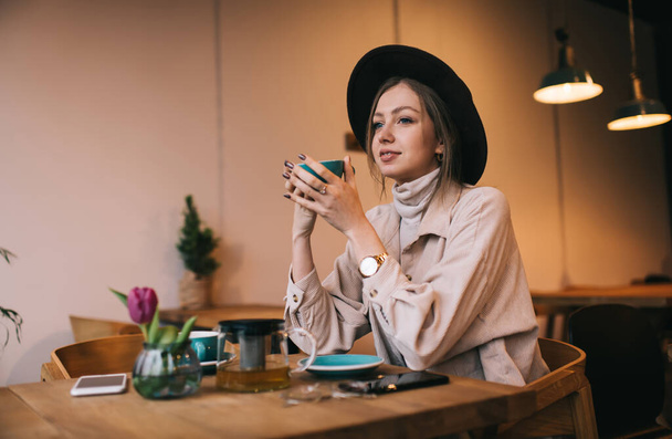 Positive lady with brown hair in trendy outfit and hat drinking hot tea and thoughtfully looking away while resting in cozy cafe - Valokuva, kuva