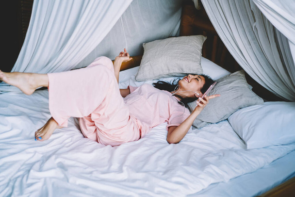 Carefree female in bedclothes enjoying morning time with perfect music browsed on smartphone device - Фото, изображение