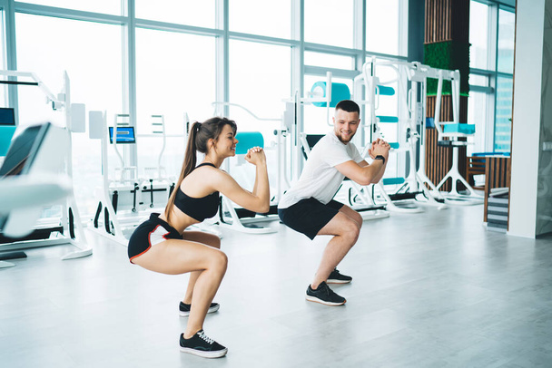 Side view of young slim sportswoman in active wear exercising with glad colleague against fitness machines near window in gymnasium - Фото, зображення
