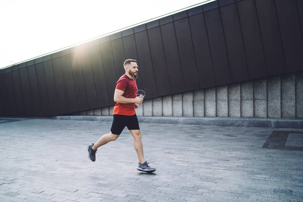 Side view of full body young sportsman running with smartphone and earphones while listening to music and looking away during morning workout - Fotó, kép