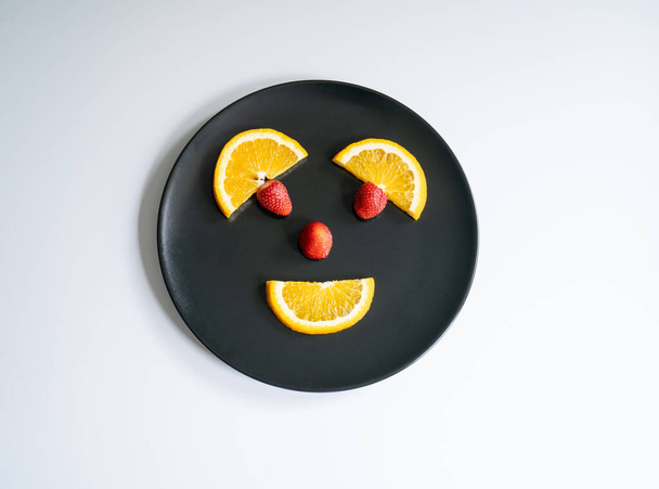 rodajas de limón y naranja, fresa en plato negro, sobre un fondo blanco. Sonríe cara  - Foto, imagen