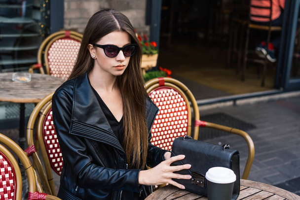 Señora de moda de confianza en chaqueta de cuero negro y gafas de sol con elegante bolso sentado a la mesa en la cafetería al aire libre con taza de papel de café - Foto, Imagen