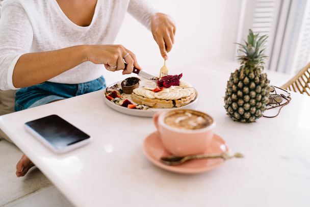 From above of anonymous female in casual clothes eating delicious breakfast with knife and fork at table with cup of coffee and pineapple - Foto, Bild