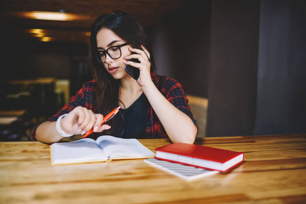 Concentrated female student in glasses and casual clothes looking at wristwatch and thinking about project while sitting at table with notebooks in cafeteria - Photo, Image