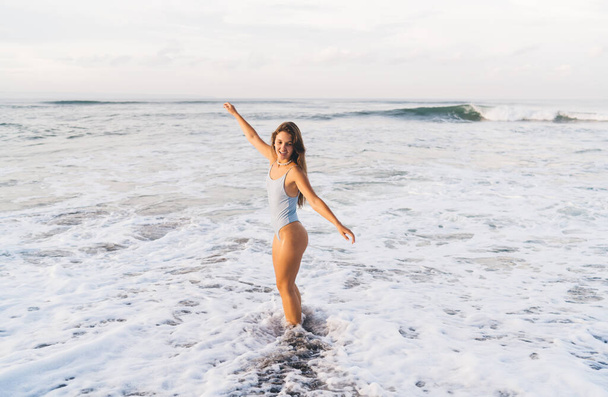 Side view full length positive slim female in white swimwear standing with arms outstretched on seashore washed by foamy sea waves - Foto, immagini