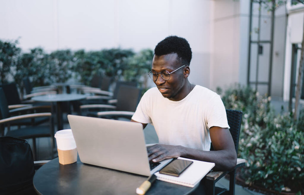 Smiling black guy in white t shirt and eyeglasses typing on laptop sitting at table with plastic cup of coffee notepad - Zdjęcie, obraz