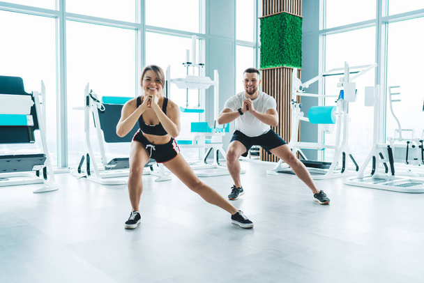 Volledig lichaam van glimlachende man en vrouw in activewear kijken naar de camera terwijl het doen van warming-up oefeningen in fitnesscentrum - Foto, afbeelding