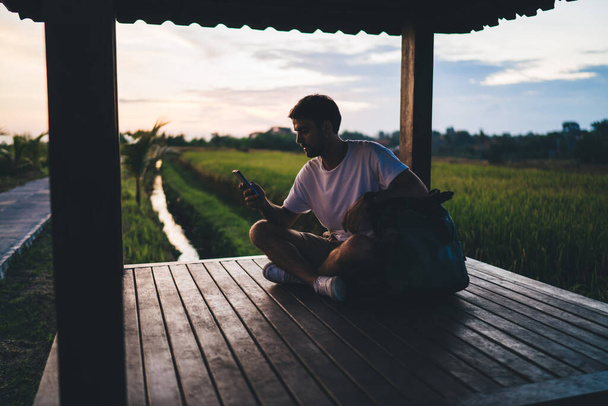 Young male traveler with backpack sitting in wooden pergola near green field and using tourist navigation app on smartphone in warm dusky evening - Valokuva, kuva