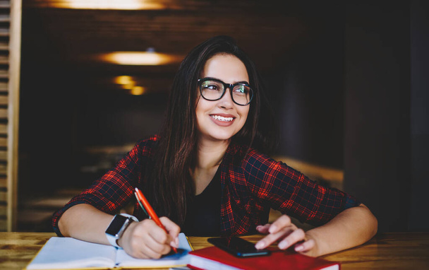 Happy female student in glasses and casual outfit taking notes in notebook and sitting at table with smartphone in cafe in daytime - Фото, изображение