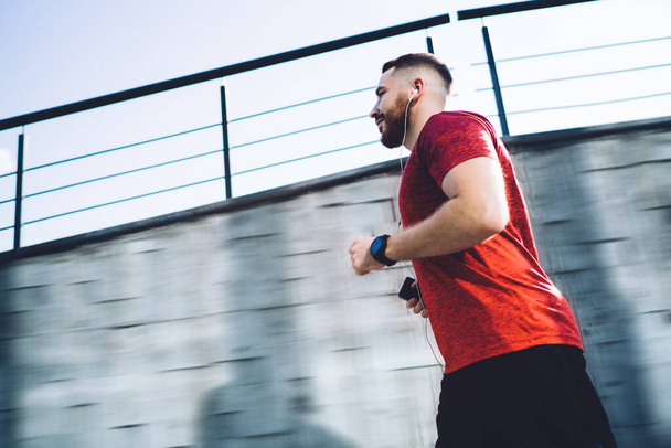 From below of confident man in sportswear looking away running and listening to music in earphones while training on street in cloudless weather - Foto, imagen