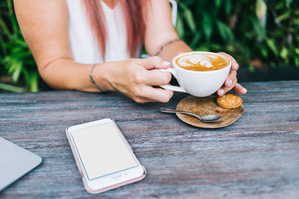 Crop unrecognizable female hipster with dyed hair sitting at summer restaurant and drinking hot cappuccino with cookie during break after work - 写真・画像