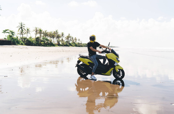 Full body of anonymous male biker in helmet sitting on scooter among wet beach while spending time in hot country and looking away - Foto, afbeelding