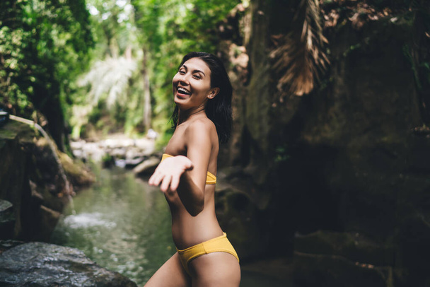 Smiling female traveler standing near stream and green tropical plants in sunny summer day wearing bikini and looking at camera - Foto, afbeelding