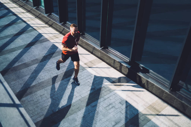 From above of full body sportsman in sneakers running with phone and earphones while training during early morning and looking away - Photo, Image