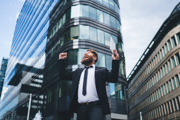 From below of bearded man in business suit looking up celebrating success and shouting on street near high modern buildings - Foto, Imagem