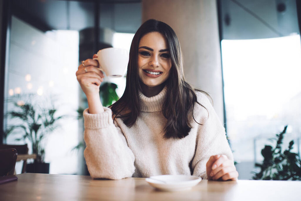 Happy female in casual outfit smiling and looking at camera while resting with cup of hot coffee and spending time in cafe - Foto, Imagem