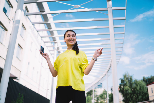From below of happy young Asian female in sporty clothes listening to music on mobile while standing on street near modern building - Фото, зображення