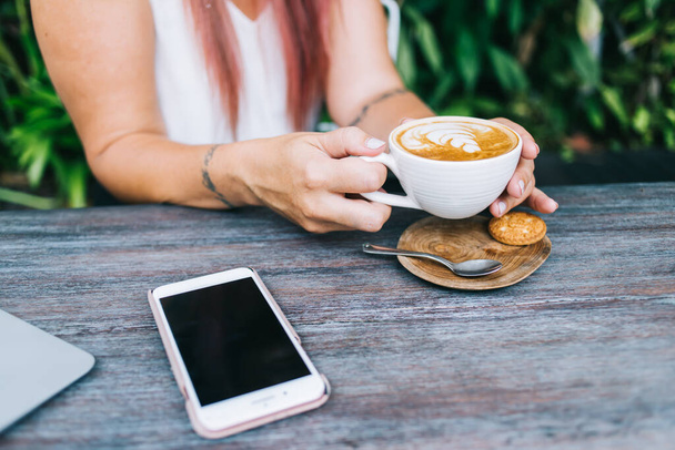 Сверху анонимная женщина в повседневной одежде сидит за деревянным столом со смартфоном и наслаждается чашкой кофе - Фото, изображение