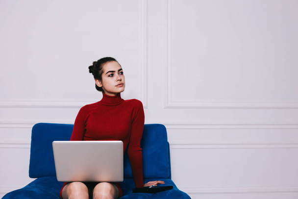 Thoughtful female freelancer in casual outfit looking away sitting on blue sofa with laptop on lap while working on project remotely - Foto, Bild