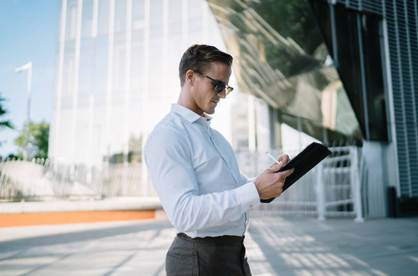 Confident fit young businessman in white shirt and dark trousers and sunglasses standing in city street and using tablet with stylus - Fotó, kép