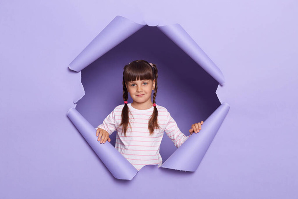 Portrait of positive happy little girl wearing striped shirt posing in torn purple paper hole, expressing optimism, looking at camera with calm expression. - Photo, Image