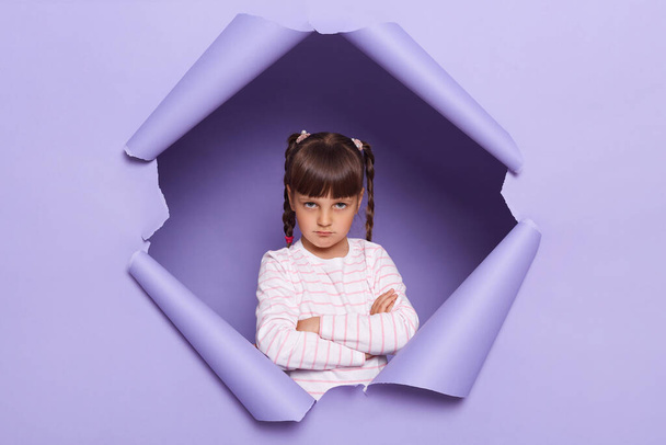 Horizontal shot of sad offended adorable little girl with braids looking through breakthrough of purple background, standing with folded hands, expressing sorrow. - Zdjęcie, obraz