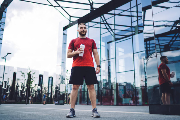 Full length of focused man in sportswear and sneakers standing on asphalt street near glass building and holding bottle of water - Fotoğraf, Görsel