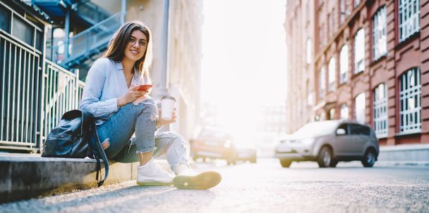  Smiling hipster blogger posing at urban street during sunny holding takeaway beverage and smartphone technology - 写真・画像