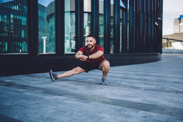 Strong male in red t shirt and black shorts looking away doing side lunge during training on street near modern building - Foto, afbeelding