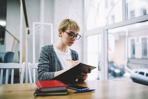 Concentrated female worker in casual clothes with eyeglasses sitting at table near window using clipboard and writing notes working in modern office - Foto, Imagem