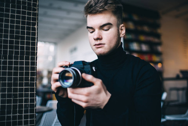 Crop pensive young man in black sweater taking photo on digital camera while standing near tile wall in library behind bookshelf - Фото, изображение