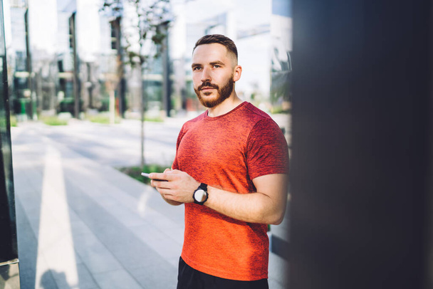 Confident man with beard standing on sidewalk near contemporary business center and text messaging on smartphone while spending time outside - Photo, image