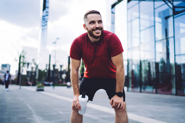 Happy fit man in sportswear with bottle of fresh water and smart watch standing on city street and resting after cardio training - 写真・画像