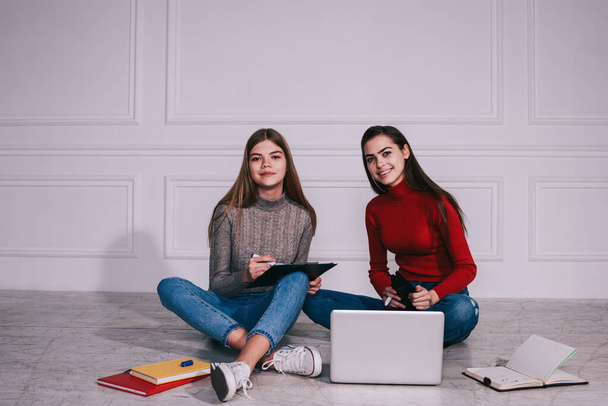 Cheerful young female students sitting on floor with crossed legs and preparing for exams using laptop and textbooks and looking at camera - Foto, Bild