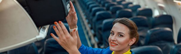 旅行スーツケースを頭上の手荷物室に置きながら航空機の旅客サロンに立つ笑顔の女性客室乗務員 - 写真・画像