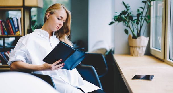 Side view of concentrated young woman in white outfit sitting in black armchair against window and taking notes in notepad - Фото, зображення
