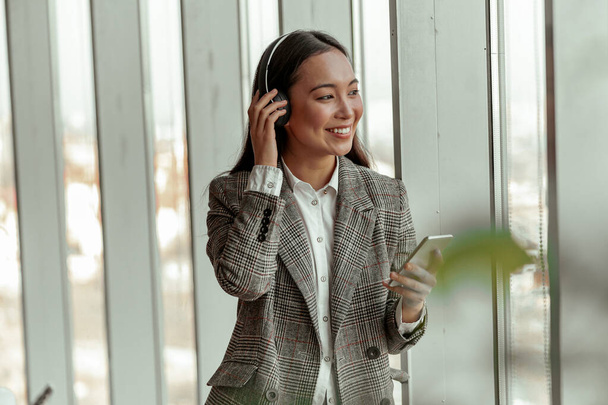 Asiática mujer de negocios escuchando música desde el teléfono en los auriculares de pie cerca de la ventana en la oficina - Foto, imagen