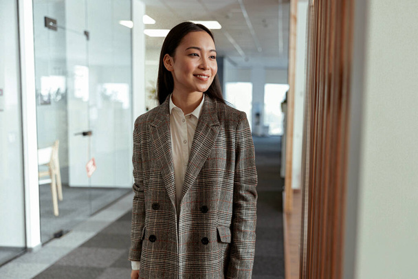Portrait of attractive asian business woman standing in modern office and looking away - Zdjęcie, obraz