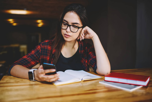 Tranquil student in glasses and casual clothes with smartphone looking at screen and working while sitting at table with books in cafe - Foto, Imagen