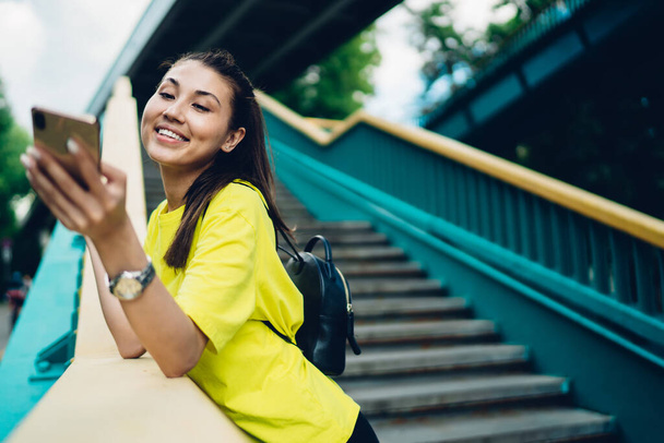Young woman with black hair in casual clothes using phone to answer SMS while walking on street in daytime and standing on stairs - Valokuva, kuva