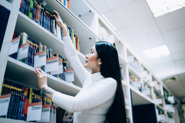 Side view from below of Asian woman in white turtle neck shirt choosing book on top shelf while standing in library - Zdjęcie, obraz