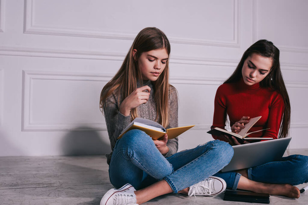 Young serious long haired female students in casual wear reading textbooks sitting on floor with legs crossed while preparing for exams - 写真・画像