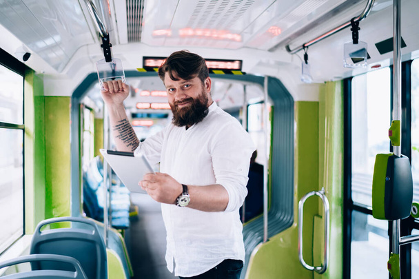 Young smiling man in casual clothes with beard standing in train and browsing tablet while looking at camera at daytime - Photo, Image