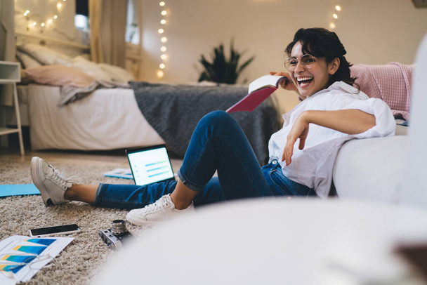 Full body side view of laughing young female freelancer sitting with netbook during remote work in cozy bedroom with glowing garlands - Фото, изображение