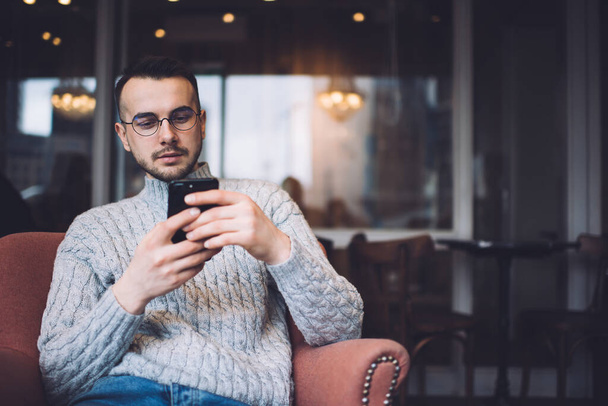 Concentrated bearded male in casual wear and eyeglasses sitting in comfortable armchair and messaging on smartphone while resting in cafe - Foto, afbeelding