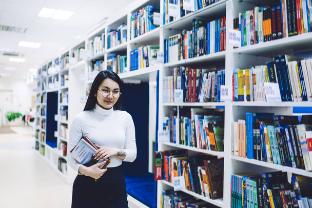 Dark haired Asian young woman with textbooks in glasses and casual clothes choosing book in bright bookstore and looking at camera - Фото, изображение