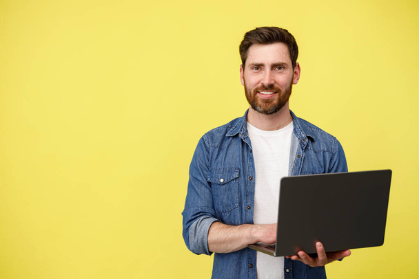 Handsome smiling man with beard holding laptop and looking camera on yellow background. Copy space - Фото, зображення
