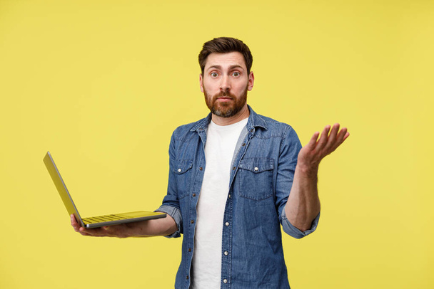 Surprised man holding a laptop and spreading his hands in confusion on yellow background - Photo, Image