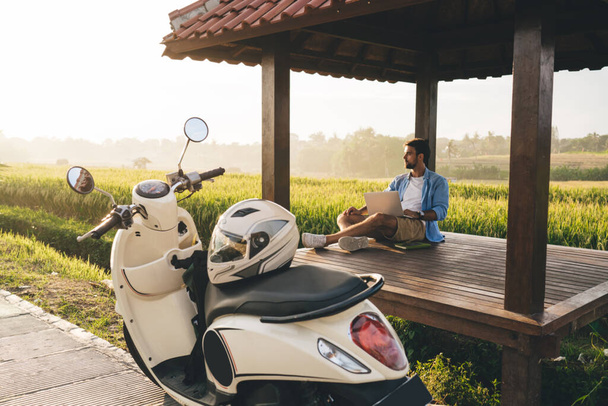 Male biker in casual wear sitting with crossed legs and netbook on wooden floor of alcove while admiring green field - 写真・画像