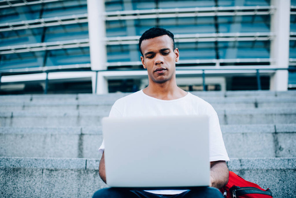 From below of serious black male freelancer in casual outfit concentrating on working on laptop remotely on stone stairs near modern building at daytime - Fotografie, Obrázek